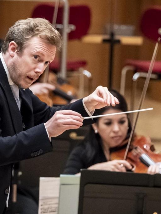 Ein dirigierender Mann steht auf einem Podest und blickt zur Seite. Vom Orchester ist nur eine Geigerin zu erkennen.