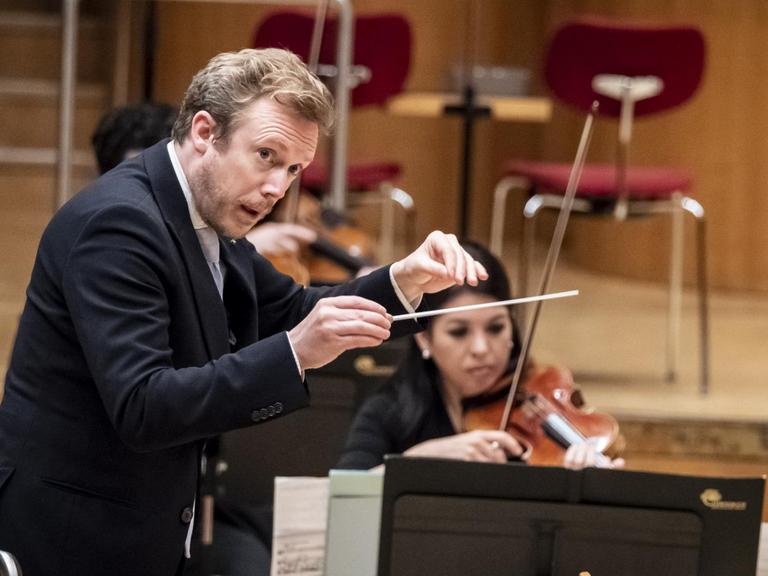Ein dirigierender Mann steht auf einem Podest und blickt zur Seite. Vom Orchester ist nur eine Geigerin zu erkennen.