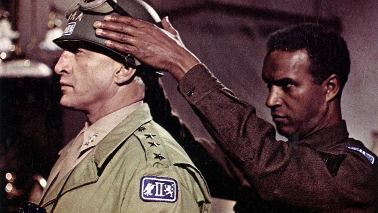 Das Standbild aus dem Film zeigt den amerikanischen Offizier Patton, der von einem anderen Offizier den Helm aufgesetzt bekommt.