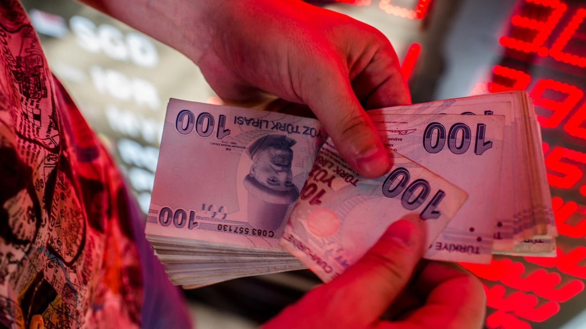 Das Bild zeigt Hände mit Banknoten der türkischen Landeswährung Lira an einem Geldautomaten. Im Hintergrund sind rot-leuchtende Wechselkurse zu sehen.