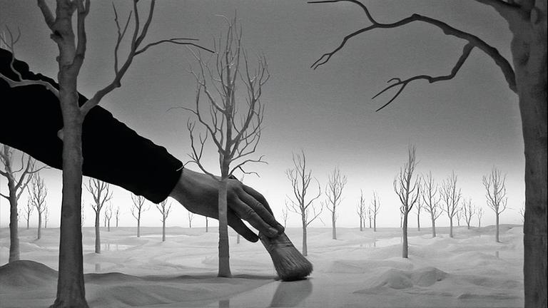 Ein Standfoto aus Hans Op de Beecks Film "Staging Silence", schwarz-weiß: Eine Hand wischt mit einem Pinsel durch eine karge Landschaft mit Bäumen.
