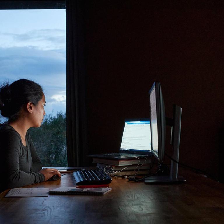 Eine junge Frau sitzt vor zwei Bildschirmen im Dämmerlicht zuhause bei der Arbeit.