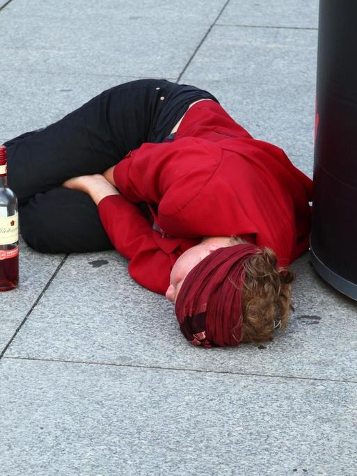 Eine junge Frau liegt völlig betrunken auf dem Pflaster an einer Plakatsäule
