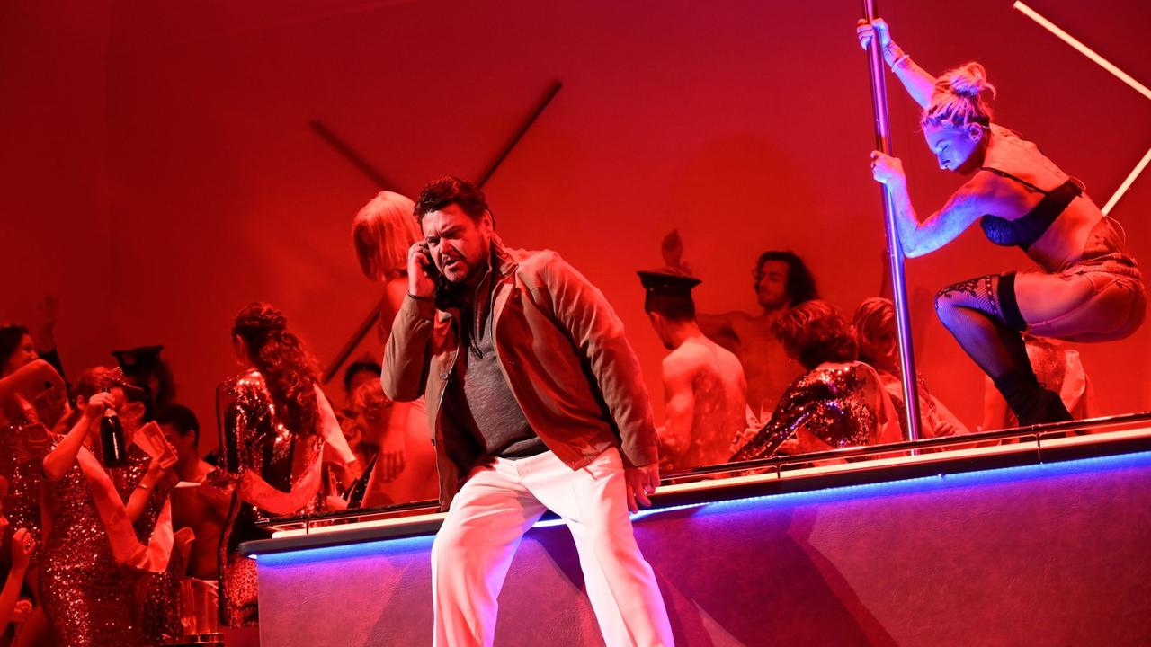 Ein Mann telefoniert zornig in einem rot ausgeleuchteten Nachtclub. Im Hintergrund tanzt eine Stripperin an der Stange.