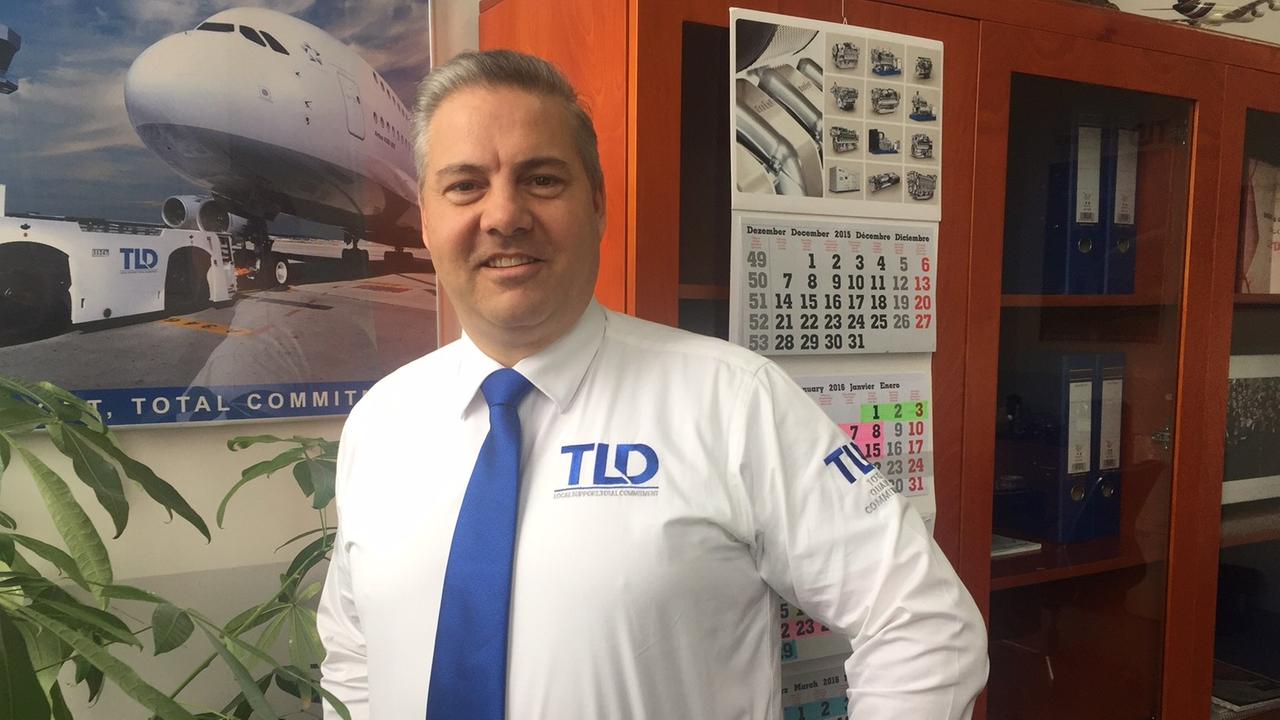 Dorn steht im weißen Firmenhemd mit blauer Krawatte in seinem Büro. Im Hintergrund sieht man einen Schrank, eine Zimmerpflanze und ein Plakat, auf dem ein Flugzeug gerade von einer TLD-Zugmaschine gezogen wird. 