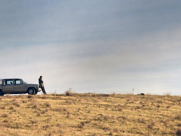 Auf einem trostlosen Hügel steht ein Mann vor einem Auto, weit entfernt von ihm steht eine junge Frau.
