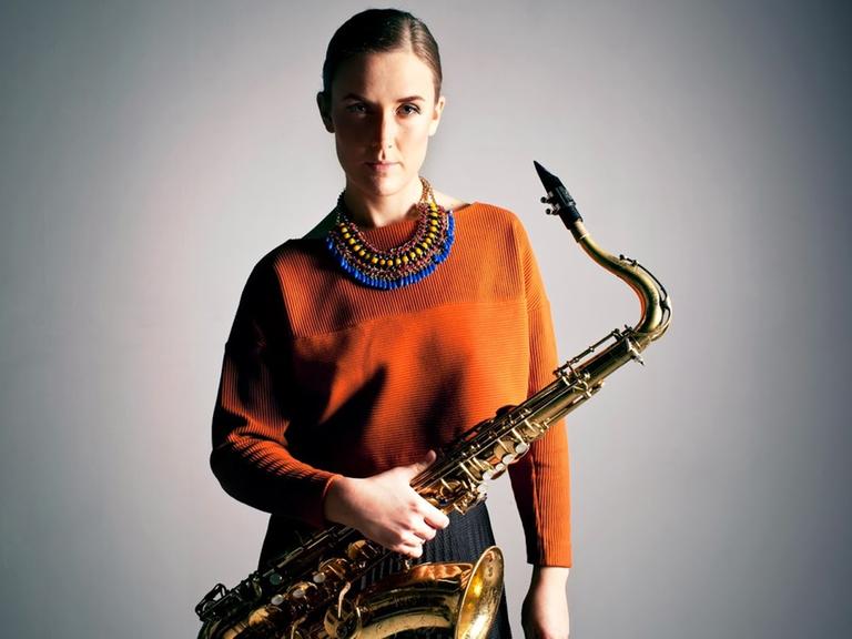 Eine Frau mit bunten Halsschmuck und Saxofon in der Hand steht im Halbschatten