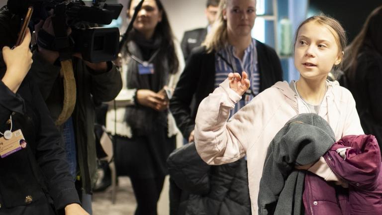 Greta Thunberg läuft umringt von Journalisten und Medientechnikern durch einen Flur und trägt ihre Jacke über den Arm geschlagen.