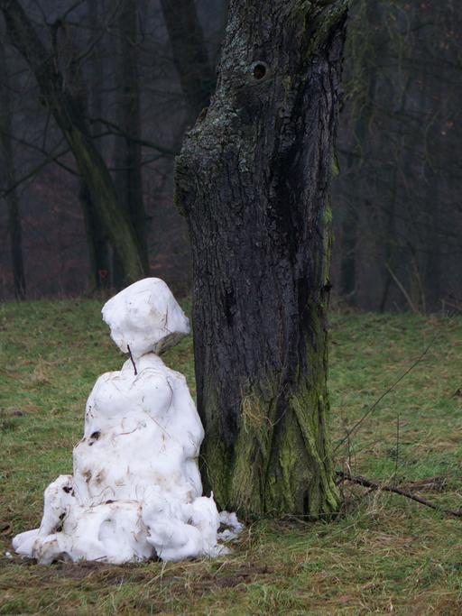 Ein dahinschmelzender Schneemann steht an einem Waldrand auf einer grünen Wiese.