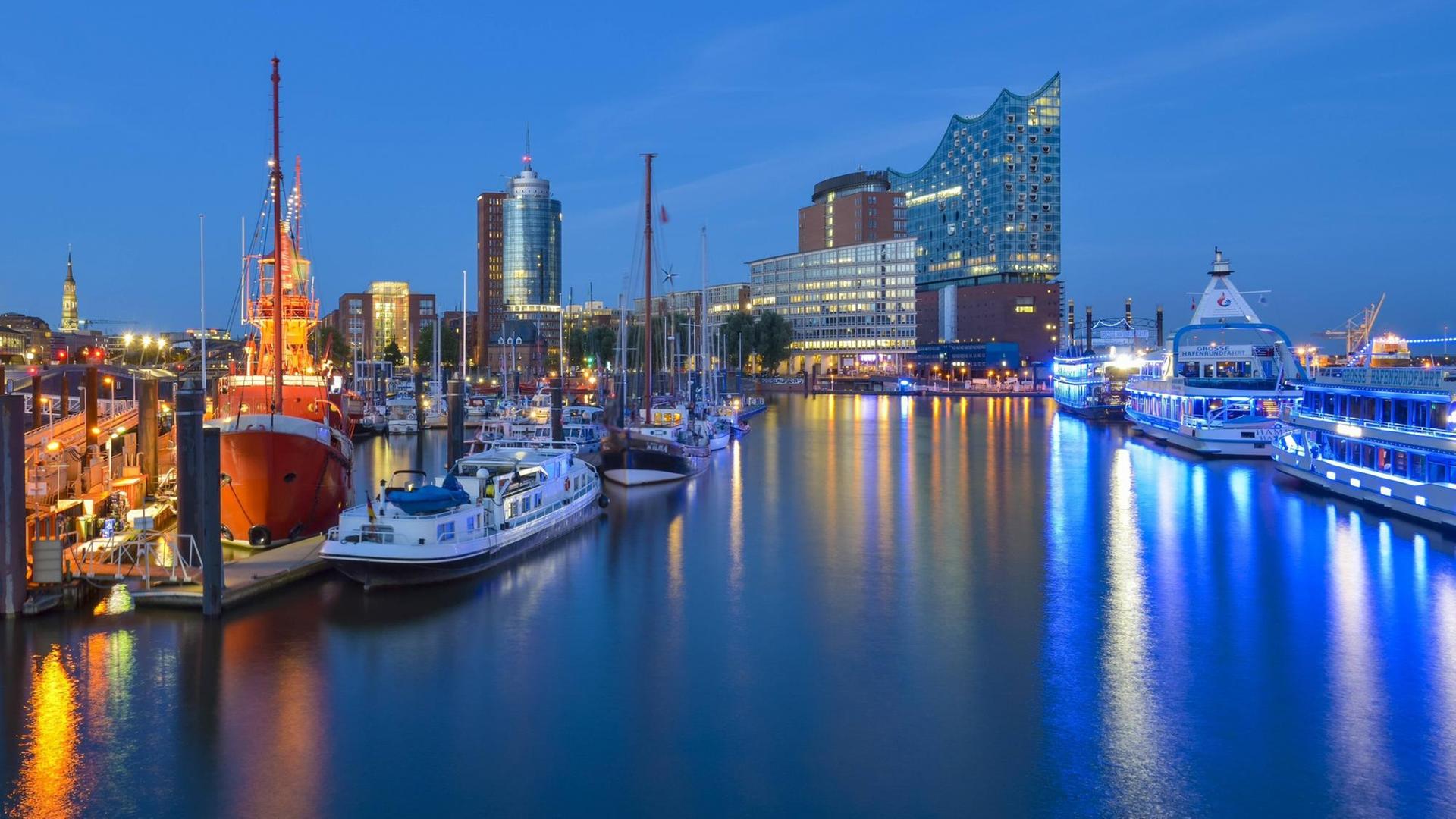 Auf dem Wasser spiegelt sich das Licht von Laternen; es dämmert. Am Horizont die Elbphilharmonie. Am linken Bilrand zwei Schiffe. Auf dem Wasser spiegelt sich das Licht von Laternen; es dämmert. Am Horizont die Elbphilharmonie. Am linken Bilrand zwei Schiffe.