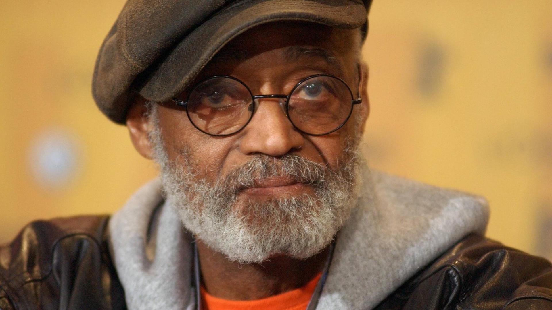 Porträt von Melvin van Peebles. Er trägt seine charakteristische Brille mit runden Gläsern, eine Schiebermütze, Lederjacke und einen grauen Kapuzenpullover.