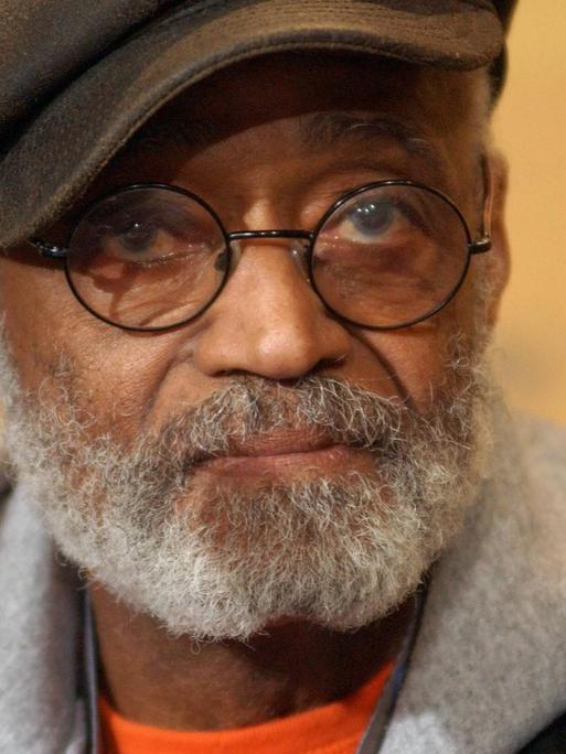 Porträt von Melvin van Peebles. Er trägt seine charakteristische Brille mit runden Gläsern, eine Schiebermütze, Lederjacke und einen grauen Kapuzenpullover.