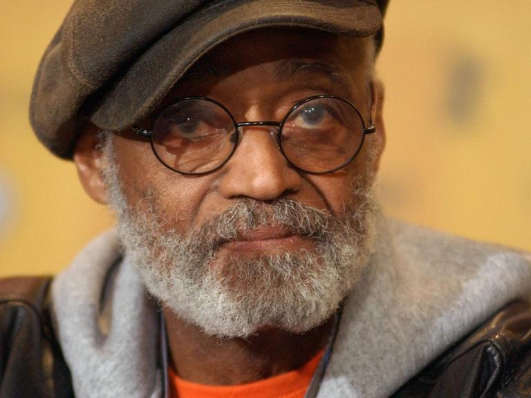 Porträt von Melvin van Peebles. Er trägt seine charakteristische Brille mit runden Gläsern, eine Schiebermütze, Lederjacke und einen grauen Kapuzenpullover.