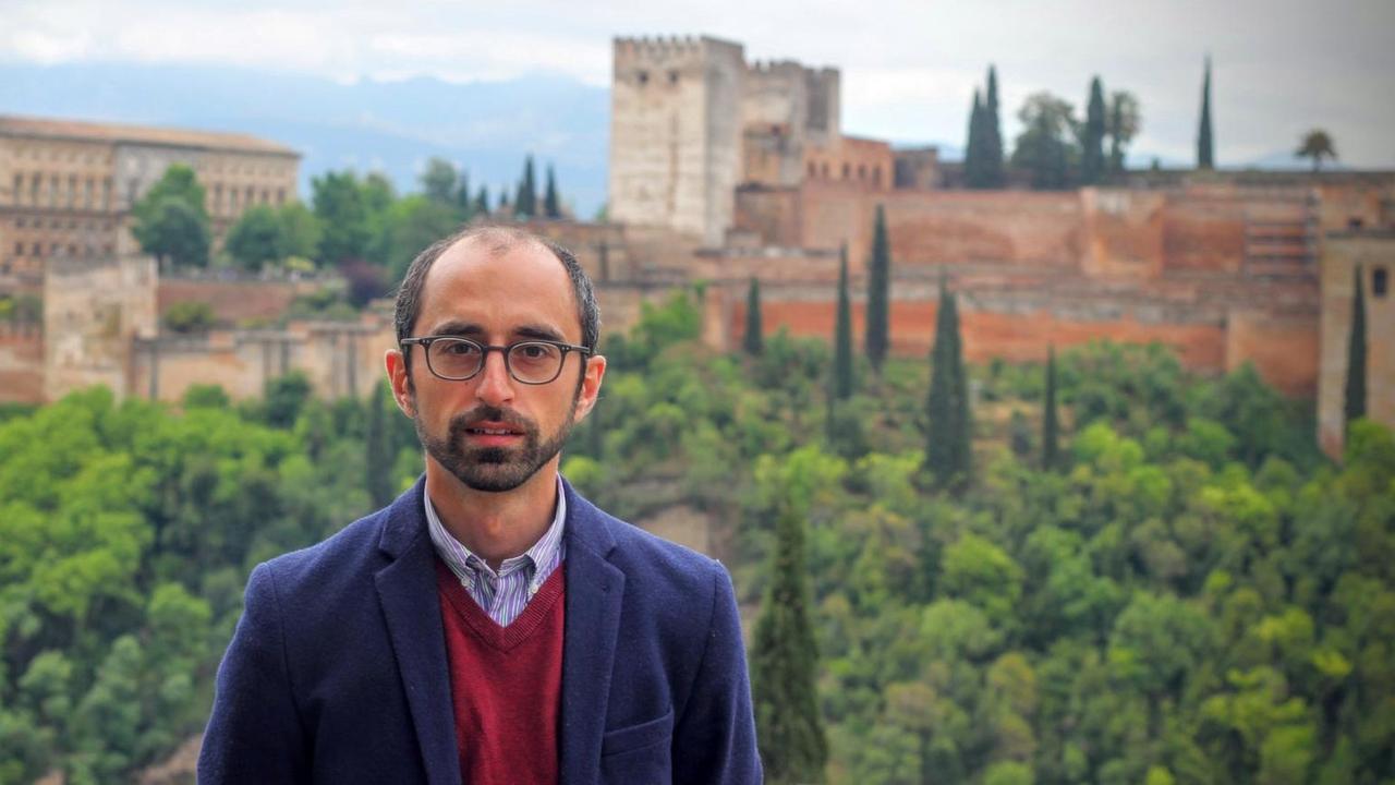 Eren Güvercin steht vor einer mediterranen Landschaft mit Zypressen und einer Festungsanlage im Hintergrund.