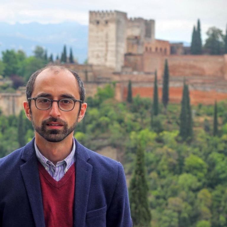 Eren Güvercin steht vor einer mediterranen Landschaft mit Zypressen und einer Festungsanlage im Hintergrund.