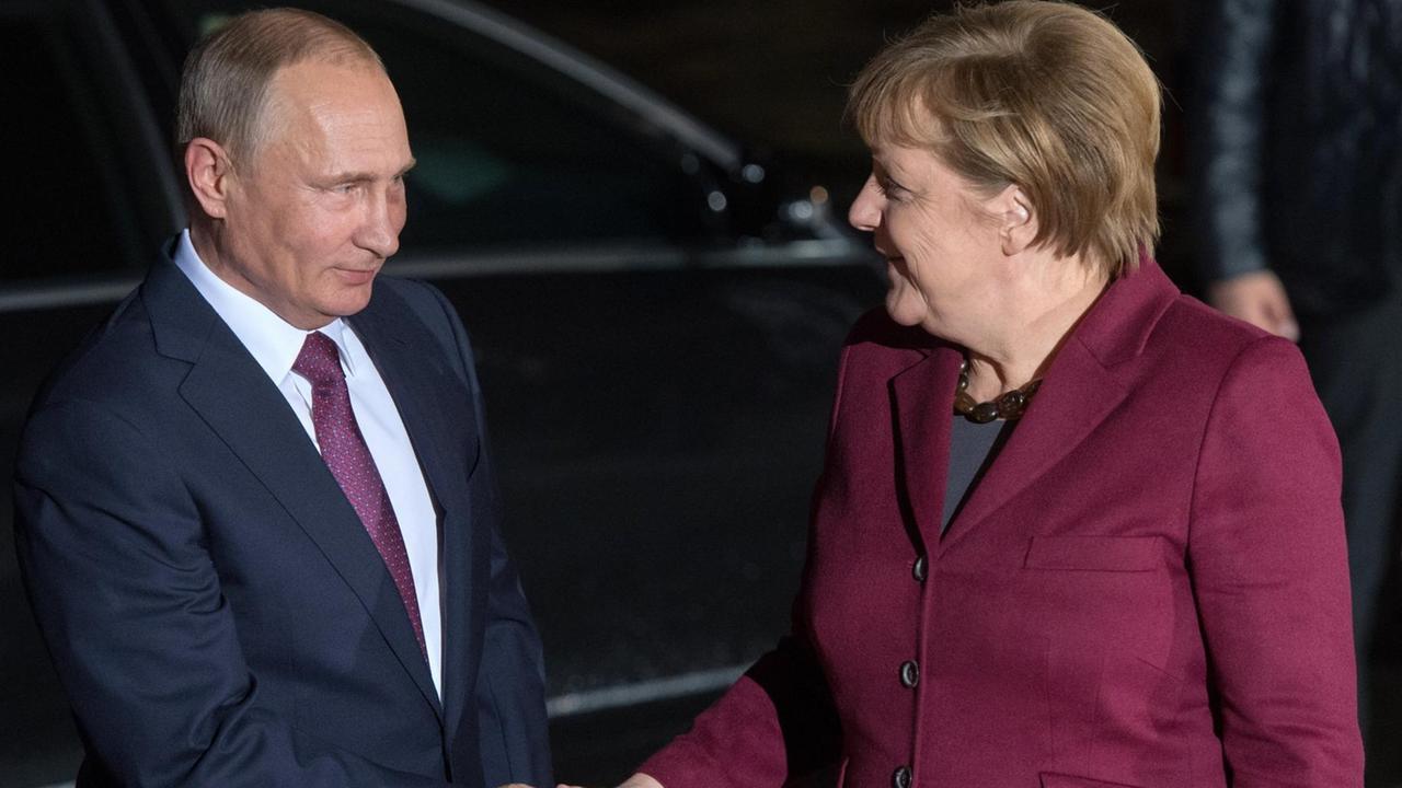 Merkel und Putin geben sich vor einem schwarzen Auto die Hand. Beide lächeln.
