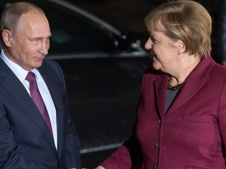 Merkel und Putin geben sich vor einem schwarzen Auto die Hand. Beide lächeln.