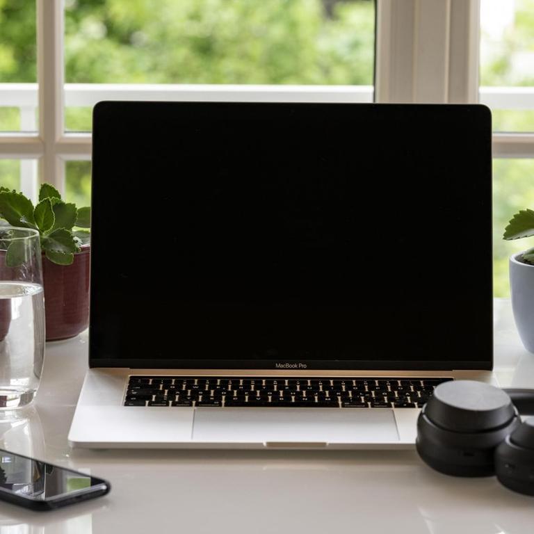 Schreibtisch mit Laptop, Telefon und mit Kopfhörern am Schreibtisch vor einem Fenster.