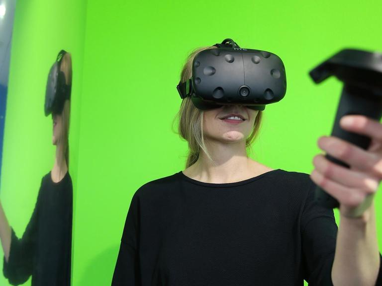 Eine Frau steht am 23.05.2017 im NRW-Forum in Düsseldorf vor einem grünem Hintergrund. Sie trägt eine Virtual-Reality-Brille und hält einen PC Controller, mit dem sie ihre virtuelle-Welt und Erlebnis steuert. Foto: David Young / dpa