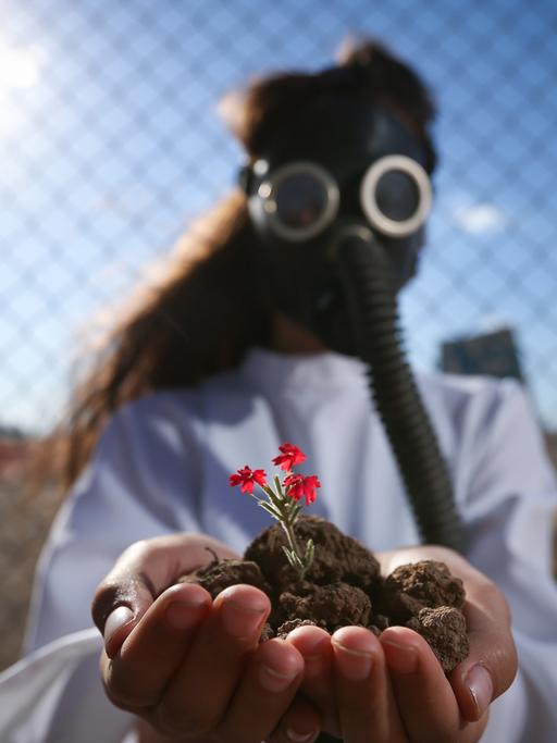 Ein Mädchen vor einem Maschendrahtzaun in einer kargen Landschaft, mit einer Gasmaske und einem weißen Hemd, hält in den göffneten Händen Erde mit einer kleinen rot blühenden Pflanze in die Kamera.