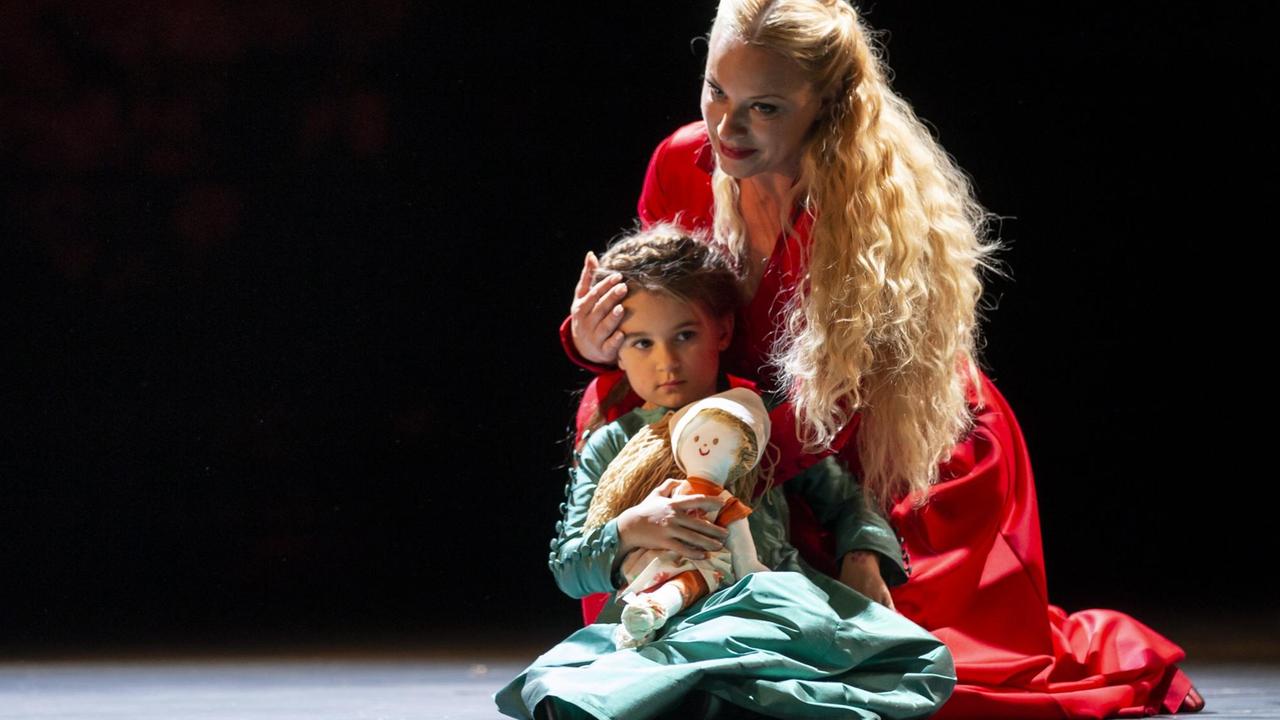 Eine Frau in rotem Kleid und langen, blonden Haaren umarmt knieend ein Kind, das wiederum eine Puppe festhält.