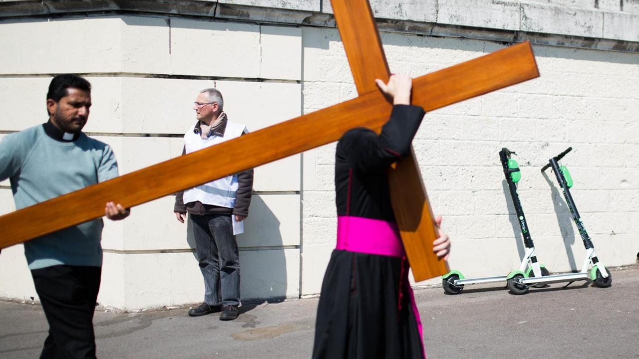 Das Foto zeigt den Pariser Bischof Denis Jachiet beim Tragen eines Holzkreuzes - im Hintergrund stehen zwei E-Scooter.