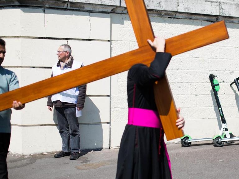 Das Foto zeigt den Pariser Bischof Denis Jachiet beim Tragen eines Holzkreuzes - im Hintergrund stehen zwei E-Scooter.