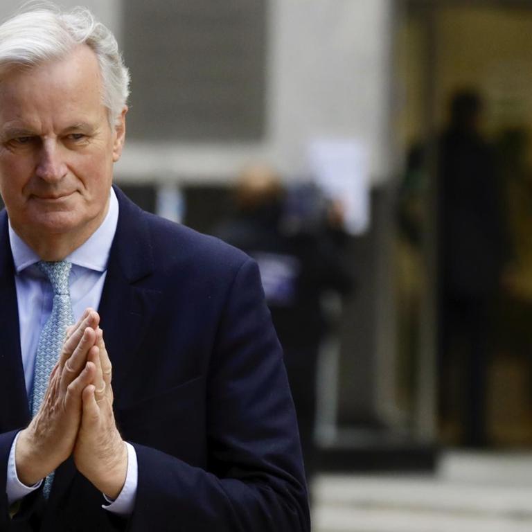 Barnier steht vor einem Eingang und hält die Hände wie beim Beten zusammen. 