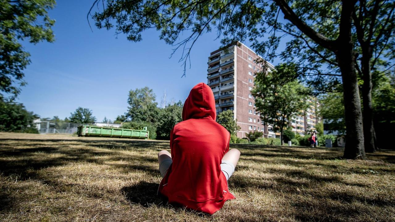 Ein jugendliches Kind sitzt in einem roten Kapuzenpullover mit dem Rücken zum Betrachter auf einer Wiese. Im Hintergrund ist ein Plattenbau zu sehen.