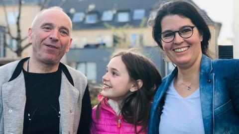 Das Foto zeigt eine lächelnde Familie bestehend aus Mutter, Vater und Tochter. Die Tochter lacht ihren Vater an.