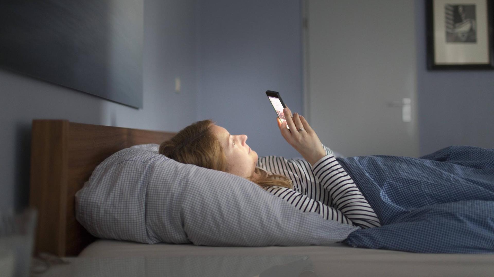 Frau liegt im Bett in einem abgedunkelten Raum und blickt auf ihr Smartphone