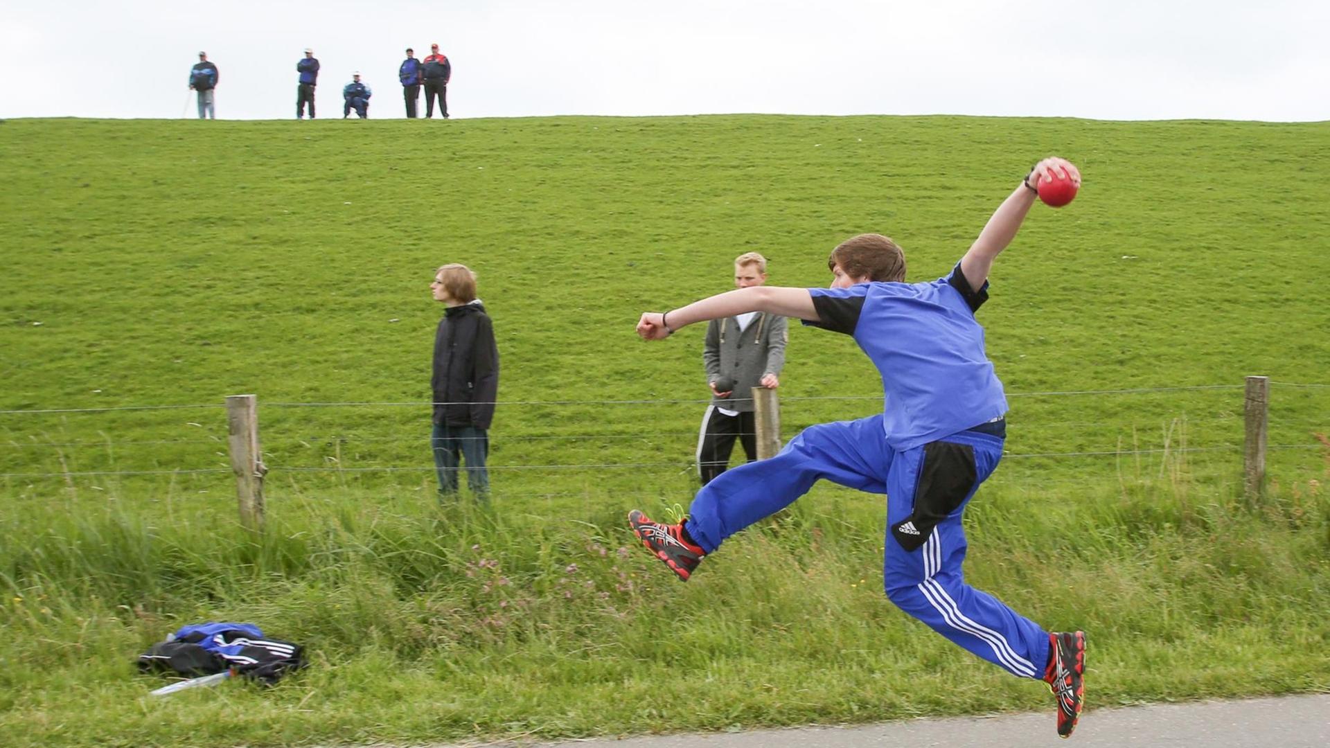 Während Zuschauer die Wettkämpfe vom Deich aus verfolgen, wirft ein Teilnehmer am 31.05.2014 bei den 8. Deutschen Meisterschaften im Boßeln in Vollerwiek (Schleswig-Holstein) eine Kugel aus Gummi.