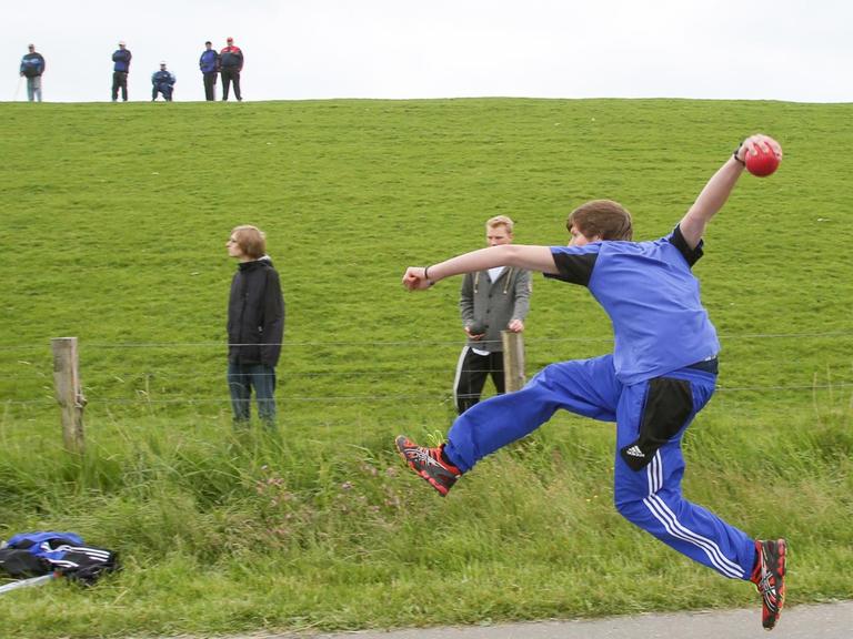 Während Zuschauer die Wettkämpfe vom Deich aus verfolgen, wirft ein Teilnehmer am 31.05.2014 bei den 8. Deutschen Meisterschaften im Boßeln in Vollerwiek (Schleswig-Holstein) eine Kugel aus Gummi.