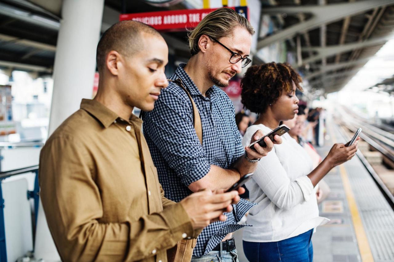 Drei Menschen stehen nebeneinander auf einem Bahnsteig und gucken auf ihre Smartphones