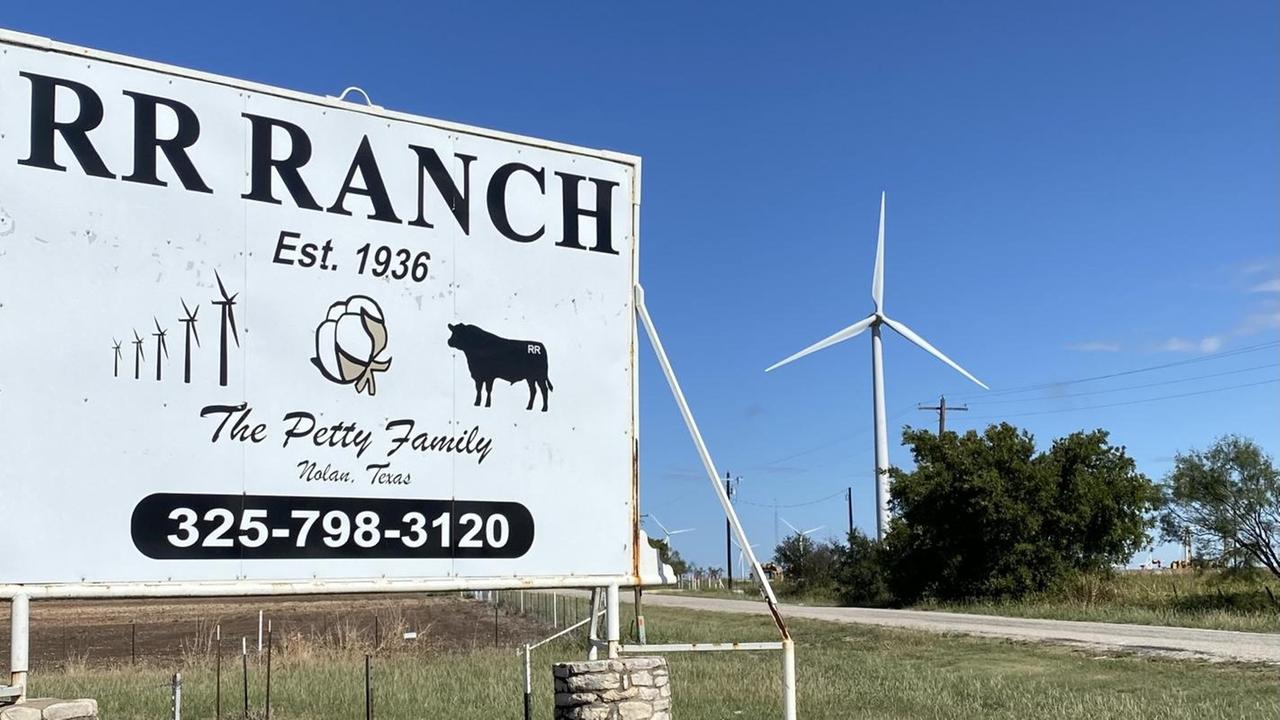 Ein großes Schild steht am Straßenrand. Darauf steht: "RR Ranch". Im Hintergrund sind Windräder zu sehen.