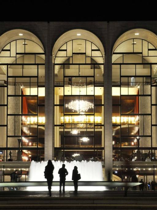 Menschen stehen im Dunklen vor einem Springbrunnen und blicken in die fünf hell erleuchteten, großen bogenförmigen Fenster des Metropolitan Opera House in New York City.