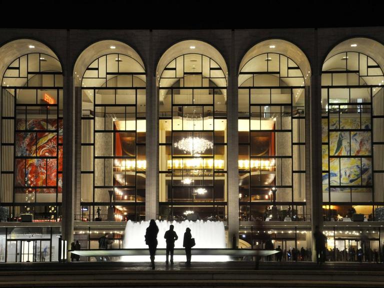 Menschen stehen im Dunklen vor einem Springbrunnen und blicken in die fünf hell erleuchteten, großen bogenförmigen Fenster des Metropolitan Opera House in New York City.