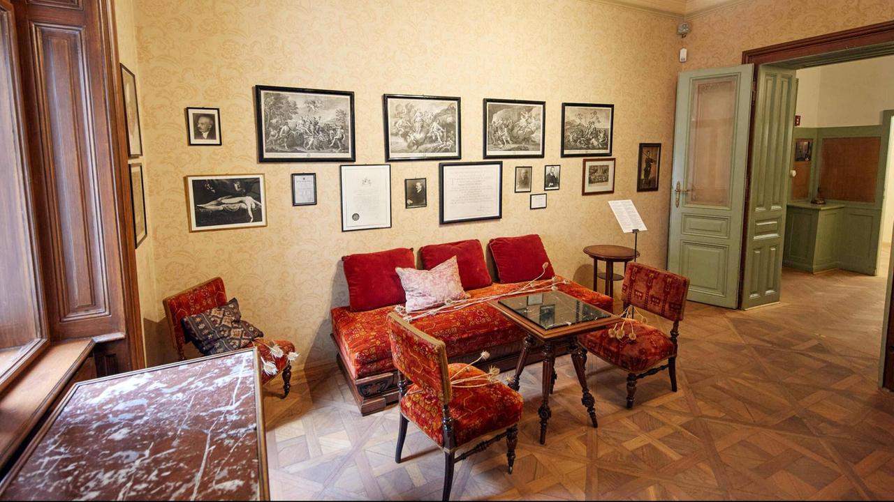 In einem Salon stehen rote Polstermöbel, an den Wänden hängen gerahmte Schwarzweiß-Bilder.