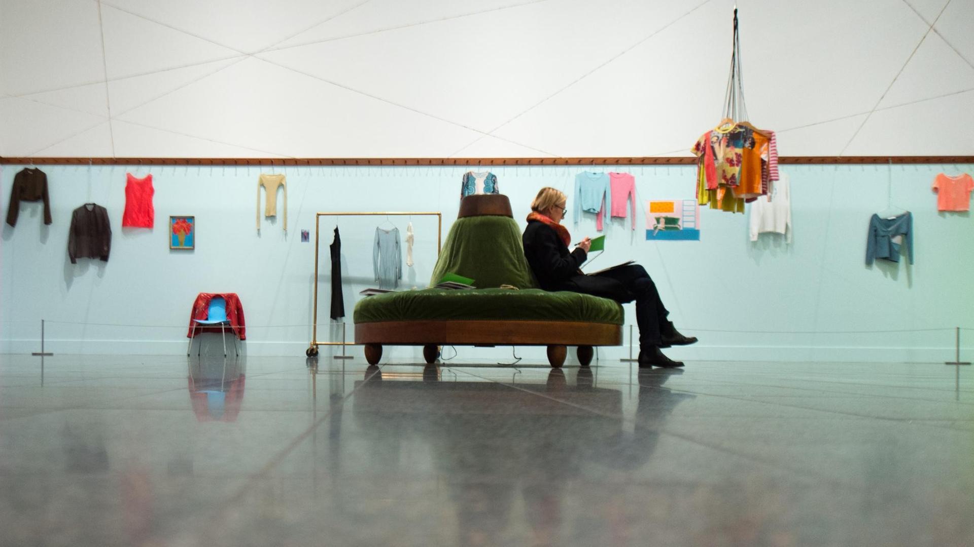 Eine Besucherin sitzt in einem Ausstellungsraum auf einem Sofa, an den Wänden hängen Kleidungsstücke.