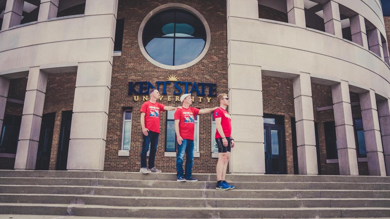 Drei Männer in roten T-Shirt stehen auf einer Treppe. Das Gebäude ist die Kent State University