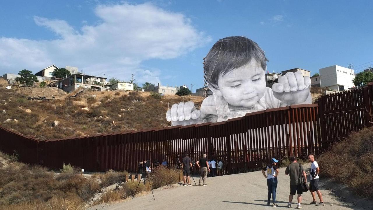 Die Installation des französischen Streetart-Künstlers JR an der Grenzmauer zwischen den USA und Mexiko: Leute stehen vor der Mauer und sehen sich das überlebensgroße Foto eines Kindes an, das über die Mauer schaut.