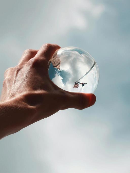 Eine Hand hält eine Glaskugel gegen den Himmel.