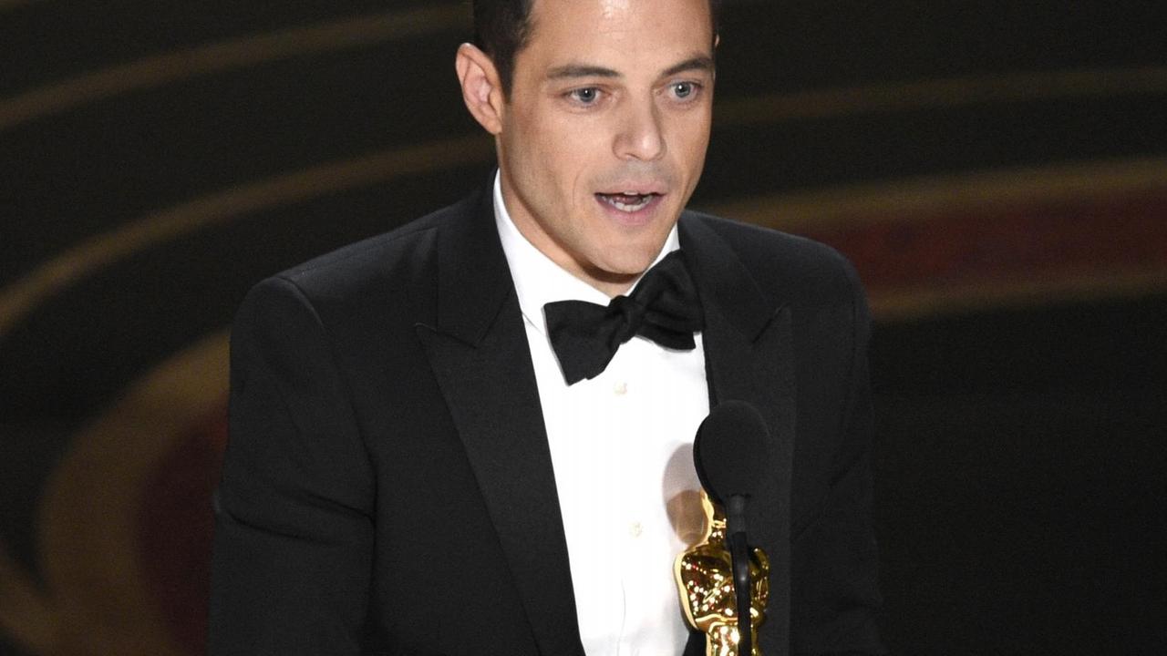Das Bild zeigt Rami Malek in einem dunklen Anzug. Er hält den Oscar in seinen Händen und spricht in ein Mikrofon. Das Bild zeigt Rami Malek in einem dunklen Anzug. Er hält den Oscar in seinen Händen und spricht in ein Mikrofon.