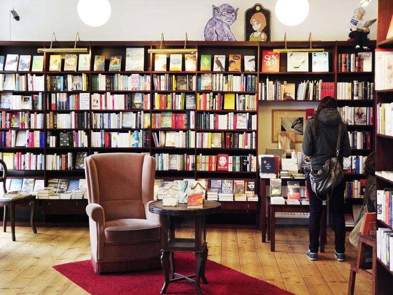 Eine Buchhandlung mit vielen Regalen. Ein Mann steht vor einem Regal und liest. In der Mitte des Raums steht ein gemütlicher Ohrensessel.