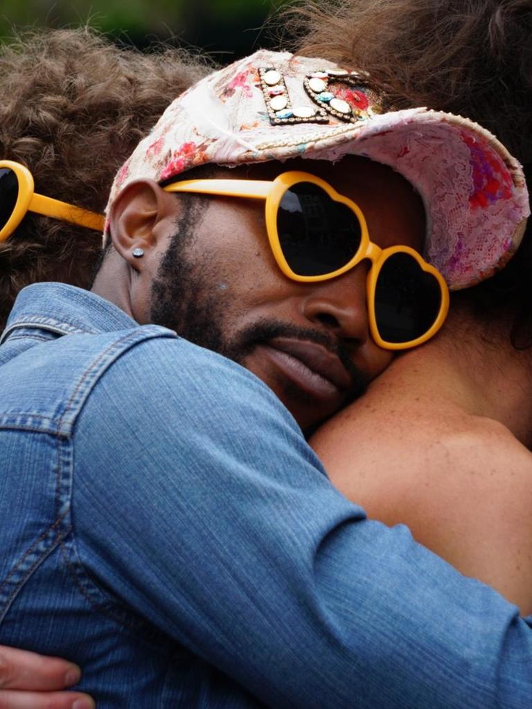 Ein Mann mit gelber Herzbrille umarmt einen anderen Mann von hinten und schmiegt sich an seinen Rücken, im Hintergrund sieht man noch mehr Menschen, alle tragen Plastikherzbrillen