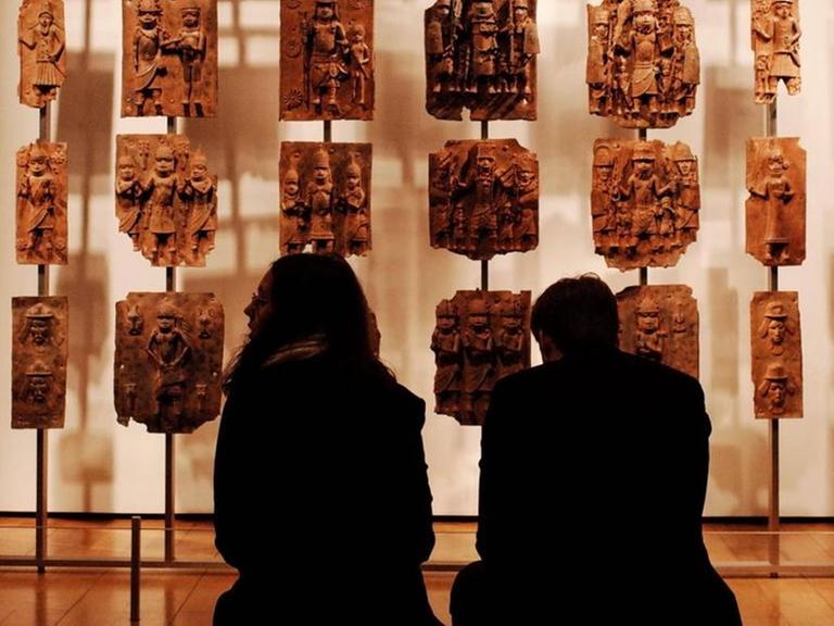 Zwei Menschen sitzen im Museum vor kunstvoll verzierten Steinplatten aus dem westafrikanischen Benin.