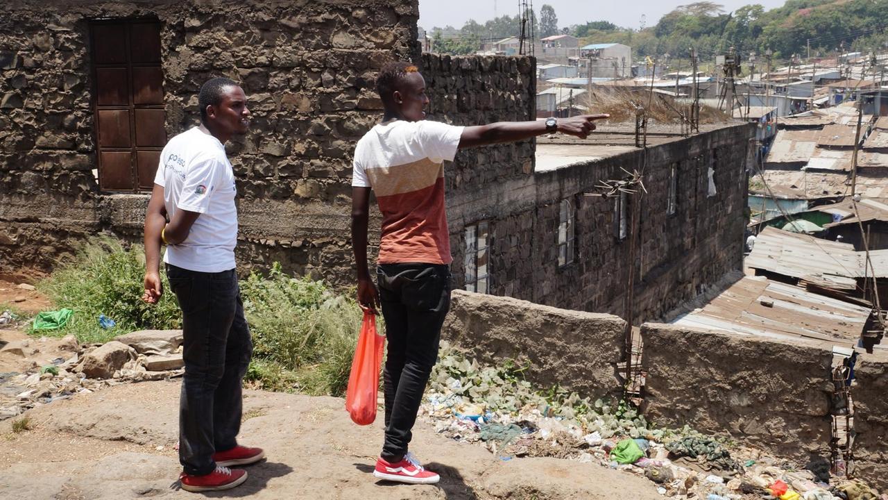 Michael Maina und Nicholas Kimeu stehen in dem Slum von Nairobi und zeigen auf die Wellblechdächer