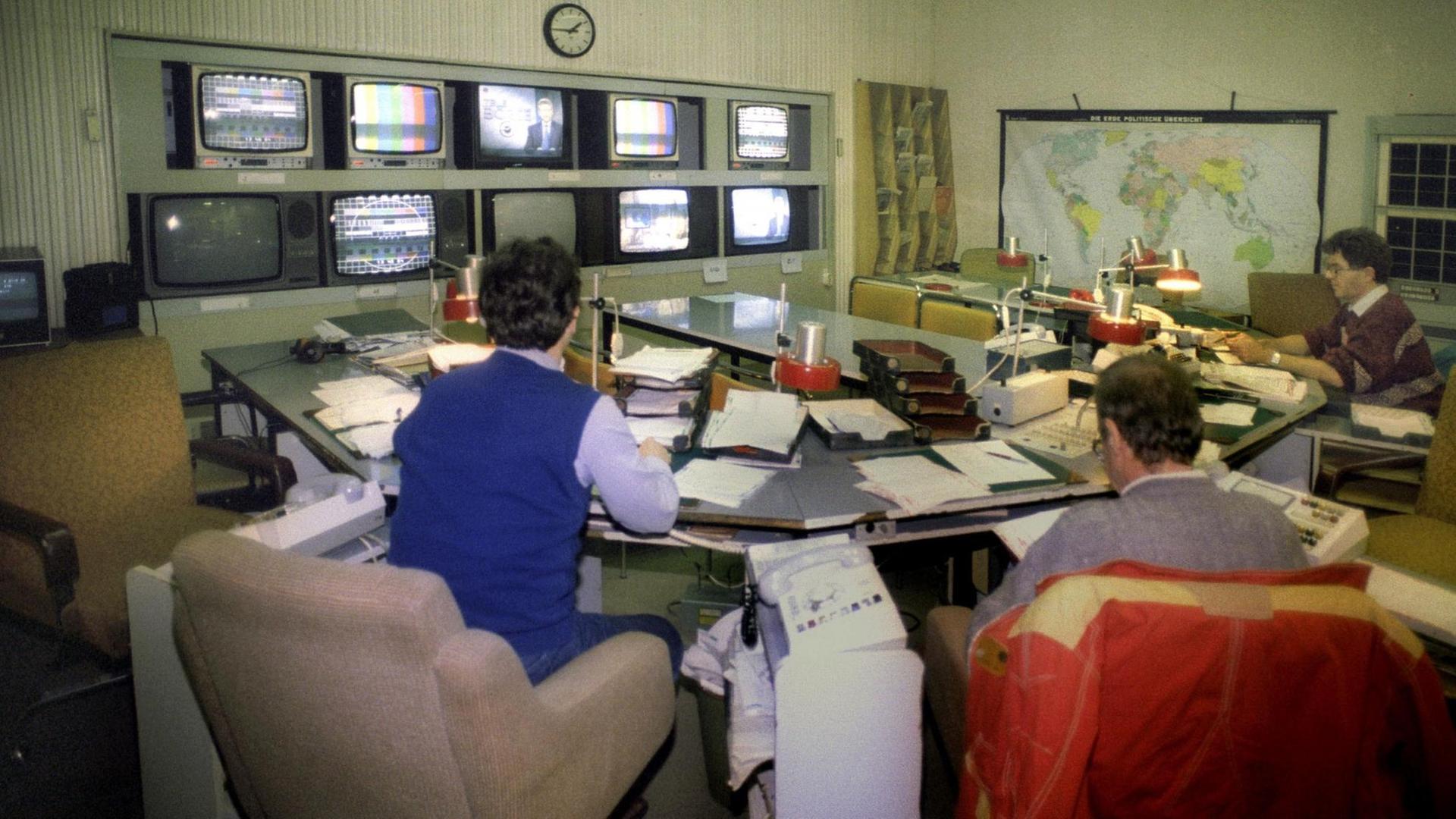 Nachrichtenraum der "Aktuelle Kamera" am 01.01.1991 in Adlershof. In einem Raum sitzen mehrere Männer, an der Wand befinden sich Monitore, neben Sesseln stehen Telefone.