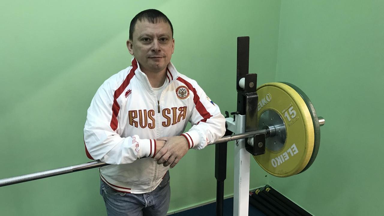 Kleiner, blasser Mann vor schweren Gewichten im Fitnesscenter in Sportjacke, auf der "Russia" steht