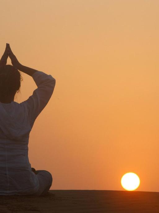 Eine Frau meditiert vor einem Sonnenuntergang.